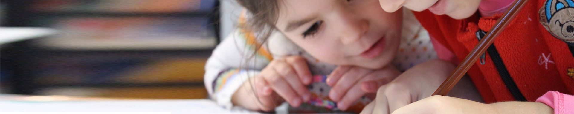 Kaksi lasta keskittyy piirtämiseen tai kirjoittamiseen pöydän ääressä. Lapsilla on värikkäät vaatteet, ja he ovat lähekkäin. Kuvan tausta on epätarkka, mutta siinä näkyy värikkäitä elementtejä.