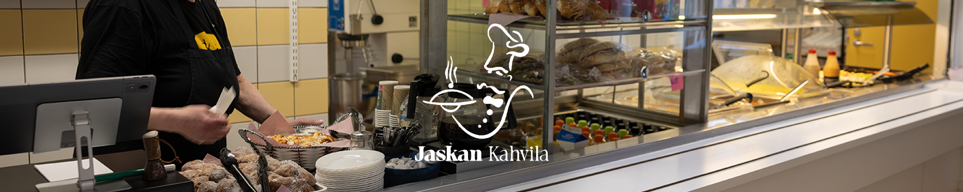 Kahvilan palvelutiski, jossa työntekijä annostelee ruokaa. Tiskillä on esillä leipiä, pullia, juomia ja muita tarjoiluja. Keskellä kuvaa näkyy ”Jaskan Kahvila” -logo.