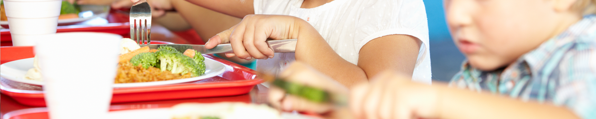 Lähikuva lasten ruokailuhetkestä, jossa näkyy kädet, punaiset tarjottimet ja lautaset, joilla on ruokaa kuten parsakaalia ja riisiä tai pastaa. Lapset käyttävät haarukoita ja veitsiä, ja taustalla näkyy valkoisia mukeja. Tilanne vaikuttaa koulun tai ruokalan ruokailuhetkeltä.