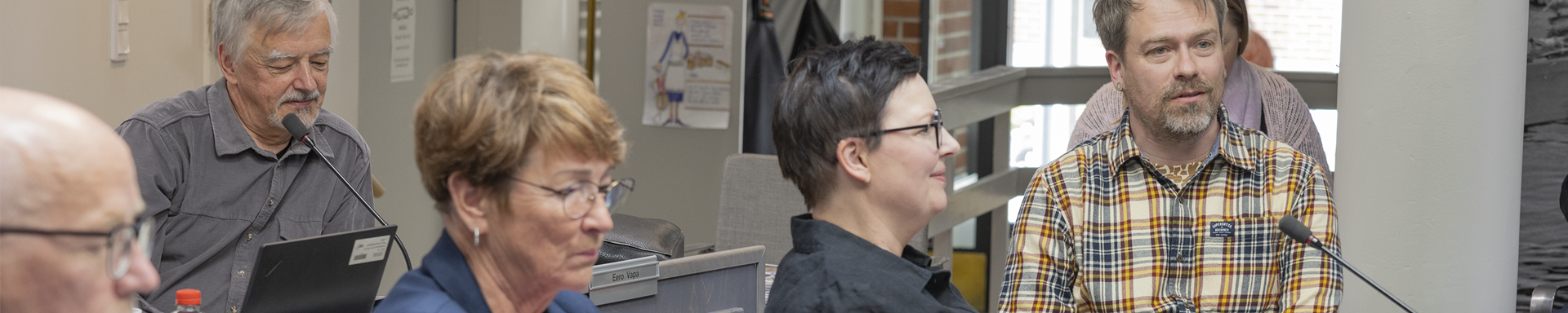 Useita henkilöitä istuu kokoushuoneessa pöytien ääressä. Etualalla näkyy kannettavia tietokoneita ja mikrofoneja. Taustalla on harmaa seinä, jossa on juliste, sekä ikkuna, josta tulee luonnonvaloa. Yksi henkilöistä käyttää ruudullista paitaa.