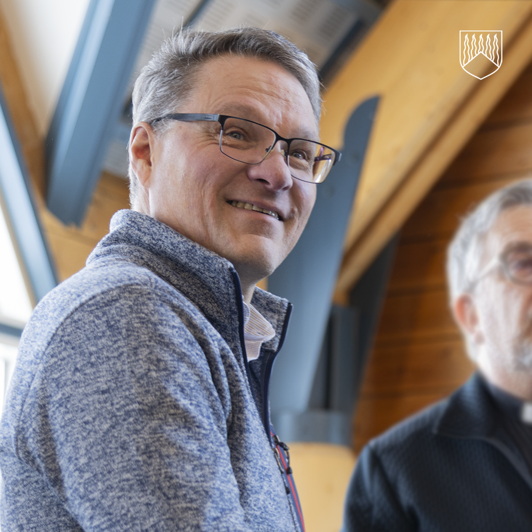 Kuvassa on sisätilassa otettu lähikuva kahdesta henkilöstä. Etualalla oleva henkilö katsoo hieman sivuun ja käyttää silmälaseja sekä vetoketjullista neuletta. Taustalla näkyy puurakenteinen tila ja kattorakenteita sekä osittain toinen henkilö. 