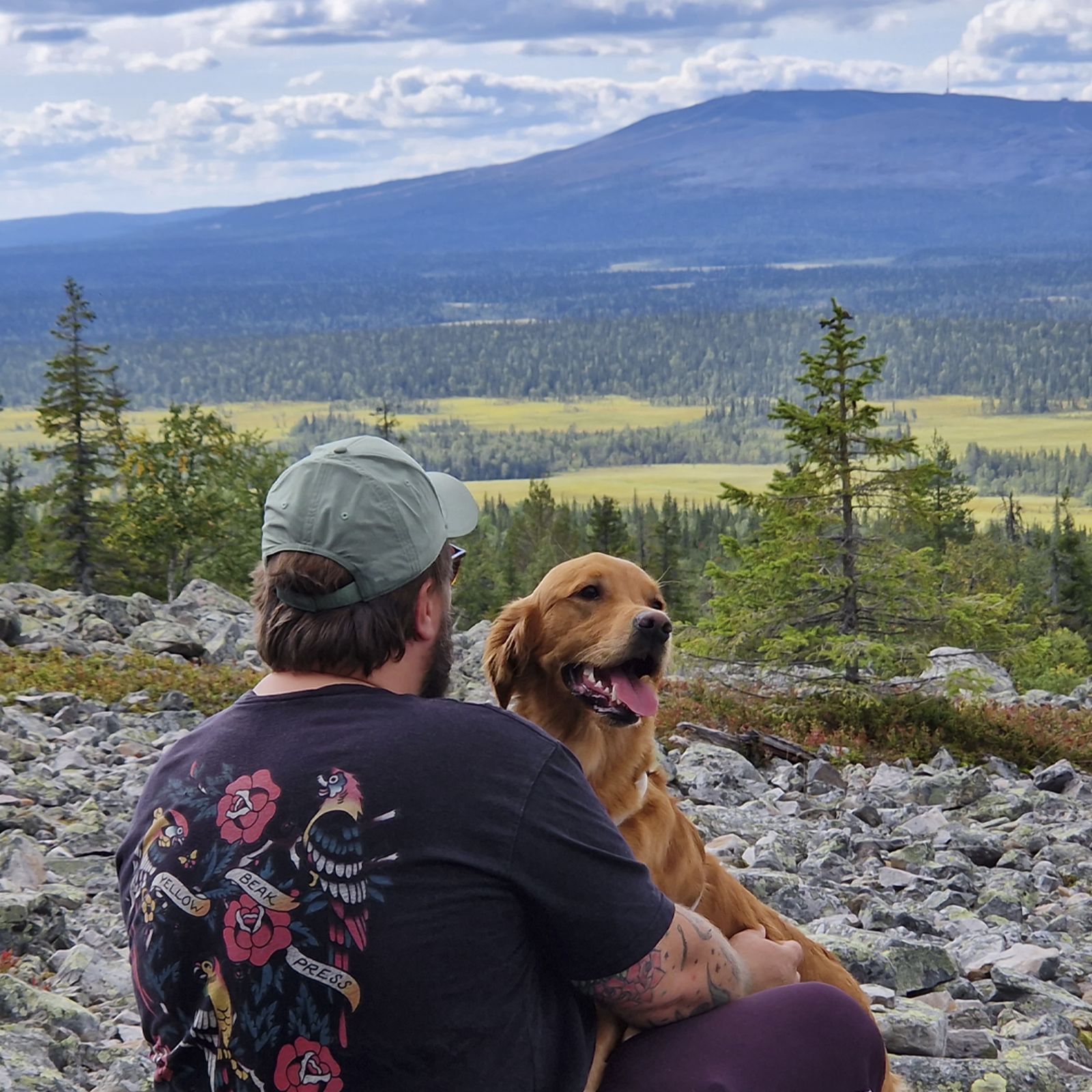 Kuvassa henkilö koiran kanssa tunturin päällä ihastelemassa tunturimaisemaa.