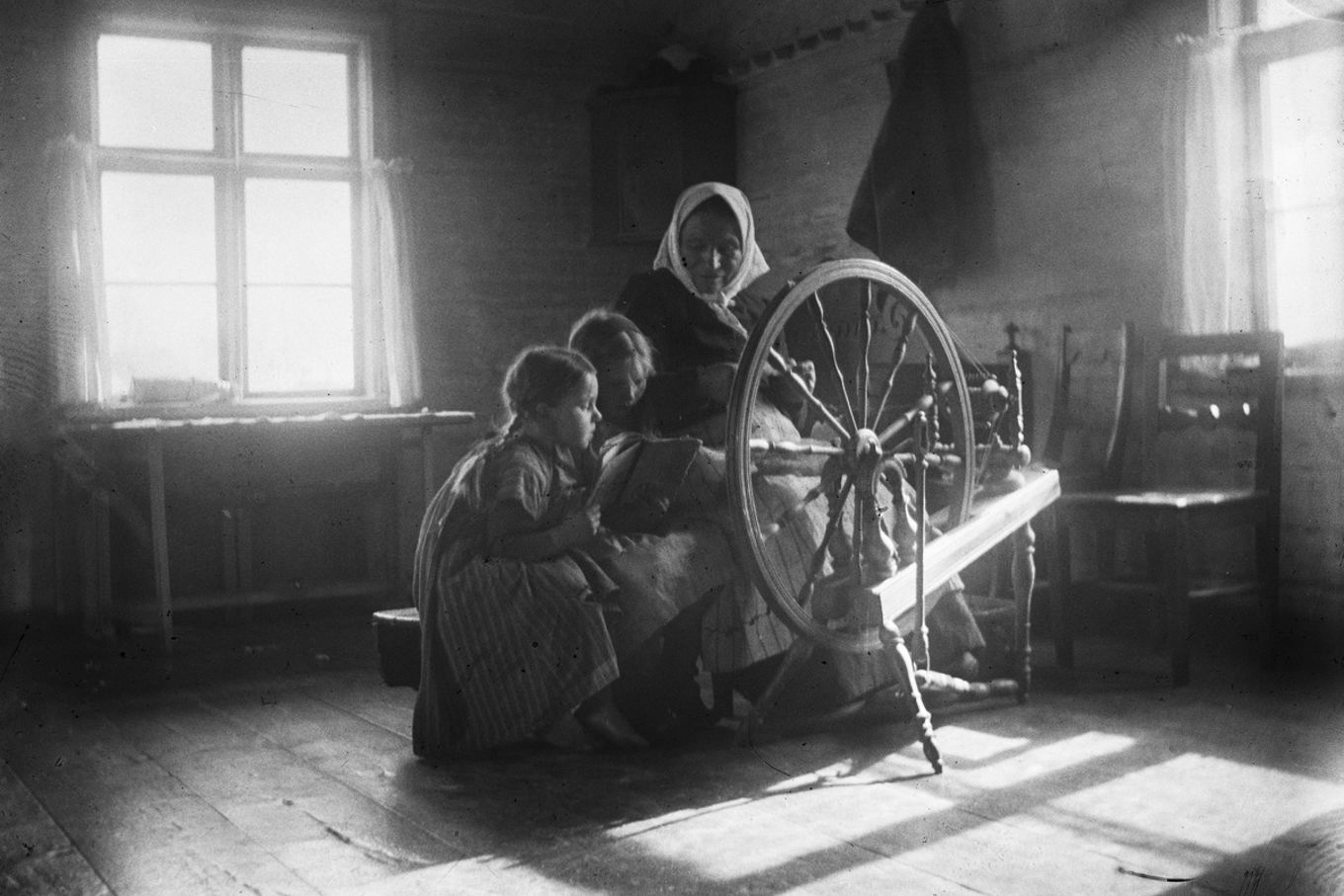 Kaksi tyttöä lukemassa huivipäisen naisen kanssa 1920-luvun Kolarissa, etualalla rukki.