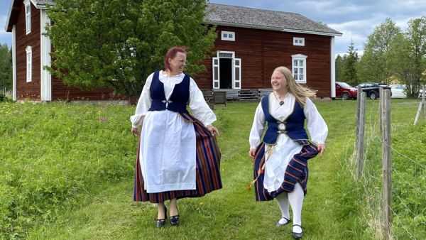 Äiti ja tytär kansallispuvuissa punamullatun talon edustalla aurinkoisena kesäpäivänä