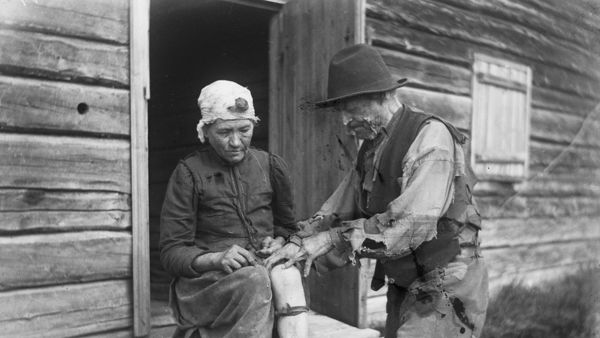 Kansanparantaja parantaa naisen kipeää polvea hirsirakennuksen edustalla 1920-luvulla