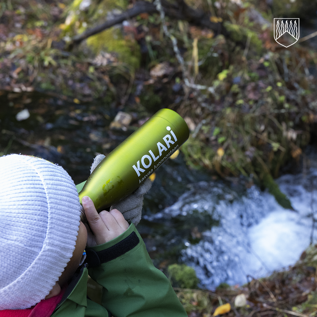 Lähikuva henkilöstä, joka juo vihreästä metallisesta juomapullosta, jossa lukee valkoisella tekstillä ”KOLARI – kasvaa luonnosta”. Henkilöllä on valkoinen neulepipo ja vihreä takki. Taustalla virtaa kirkas puro kivien ja sammaleen ympäröimänä, ja ympärillä näkyy syksyisiä lehtiä ja metsän kasvillisuutta.