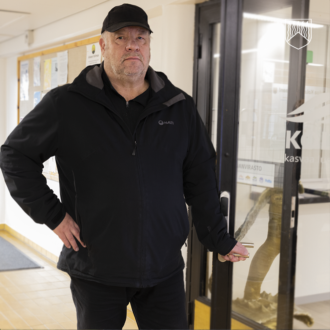 Henkilö seisoo sisätilassa lähellä lasiovea, joka johtaa huoneeseen, jossa näkyy tekstiä ja logo. Henkilöllä on päällään musta takki, musta lippalakki ja tummat housut. Vasemmassa kädessä on avainnippu, ja oikea käsi on lantiolla. Taustalla on ilmoitustaulu, jossa on kiinnitettyjä papereita, sekä vaalea käytävä.