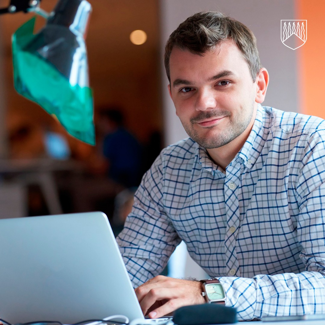 Henkilö istuu sisätilassa pöydän ääressä kannettavan tietokoneen edessä. Henkilöllä on ruudullinen kauluspaita ja ranteessa älykello. Kuvassa näkyy myös vihreä lamppu vasemmalla ja taustalla epätarkka toimistoympäristö.