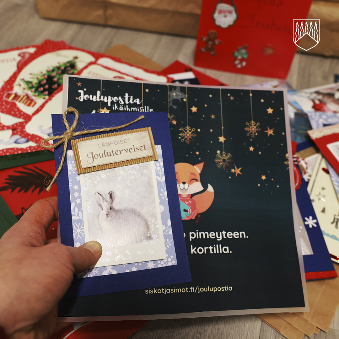 Lähikuva kädestä, joka pitelee sinistä joulukorttia, jossa on valkoinen jänis ja teksti "Jouluterveiset". Kortti on sidottu narulla. Taustalla näkyy muita joulukortteja, joista osa on punaisia ja osa sinisiä, sekä mainoslehtinen, jossa on teksti "Joulupostia ikäihmisille" ja osoite "siskotjasimot.fi/joulupostia".
