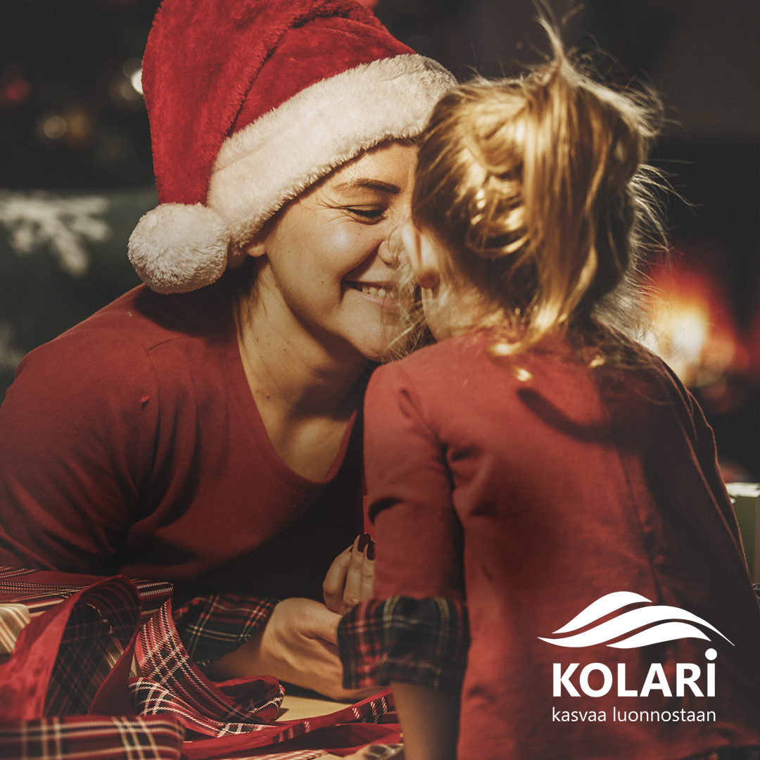 Joulutunnelmainen kuva, jossa kaksi henkilöä istuu yhdessä pöydän ääressä. Etualalla näkyy punainen ruudullinen kangas ja lahjapaperia. Vasemmalla oleva henkilö käyttää punaista jouluhattua, ja oikealla oleva henkilö on pukeutunut punaiseen asuun.