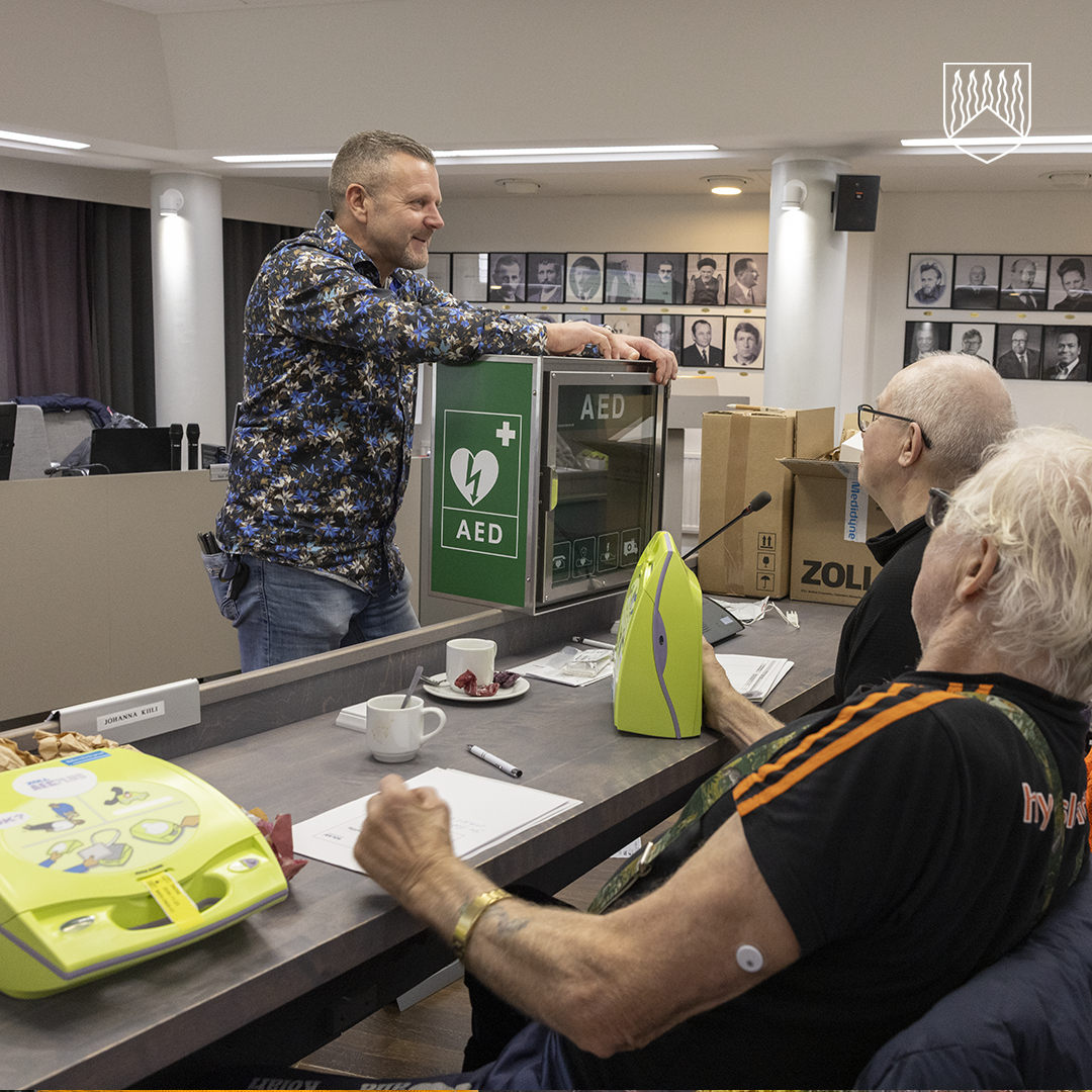 Henkilö seisoo pöydän vieressä ja esittelee vihreää seinään kiinnitettyä AED-laitetta (defibrillaattoria). Pöydällä on kaksi kannettavaa defibrillaattoria, papereita, kyniä ja kahvikuppeja. Taustalla näkyy seinällä riveittäin mustavalkoisia muotokuvia.