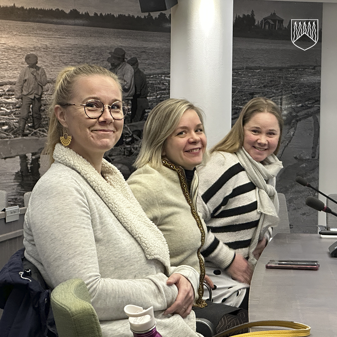 Kolme henkilöä istuu vierekkäin neuvotteluhuoneessa pöydän ääressä. Pöydällä on mikrofoneja ja vesipullo. Taustalla on suuri mustavalkoinen valokuva, jossa näkyy miehiä työskentelemässä puutavaran parissa veden äärellä. Seinällä on myös vaakuna, jossa on viisi pystysuuntaista viivaa.