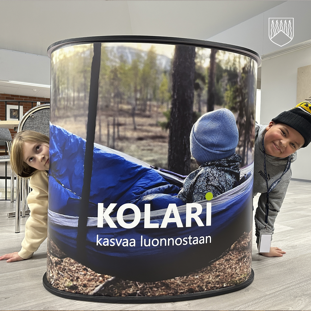 Messuosaston tai mainostelineen pinta, jossa näkyy kuva lapsesta istumassa sinisessä riippumatossa metsämaisemassa. Taustalla havupuita ja luonnonvaloa. Etualalla teksti 'KOLARI kasvaa luonnostaan' sekä Kolari-kunnan tunnus oikeassa yläkulmassa.