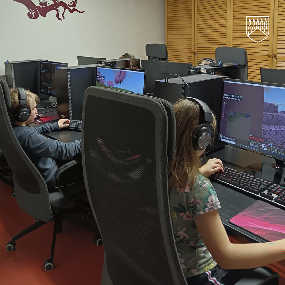 Rivissä olevat tietokoneet, joiden ääressä istuu henkilöitä pelaten videopeliä. Näytöillä näkyy Minecraft-pelin maisemia ja käyttöliittymä. Jokaisella pelaajalla on kuulokkeet ja edessä näppäimistö sekä hiiri. Huoneessa on punainen lattia, vaaleat seinät ja puinen kaapisto taustalla. Seinällä näkyy punainen koristekuvio.