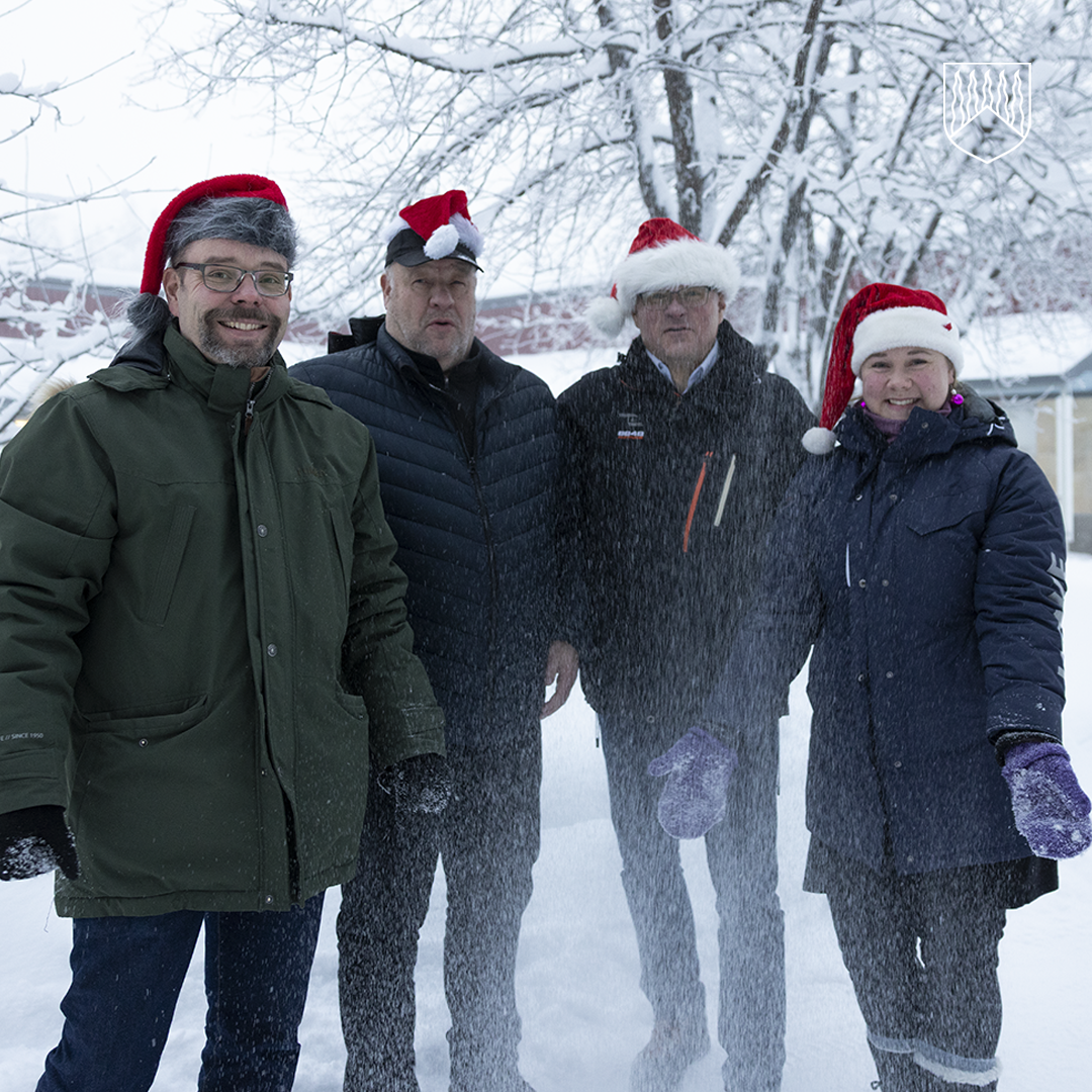 Neljä henkilöä seisoo lumisessa ulkoilmassa, taustalla lumisia puita ja rakennuksia. Kaikilla on päässään punainen joulupukin hattu ja talvivaatteet, kuten paksut takit ja hanskat. Etualalla näkyy lumisadetta, joka peittää osittain henkilöiden vaatteita ja jalkoja.