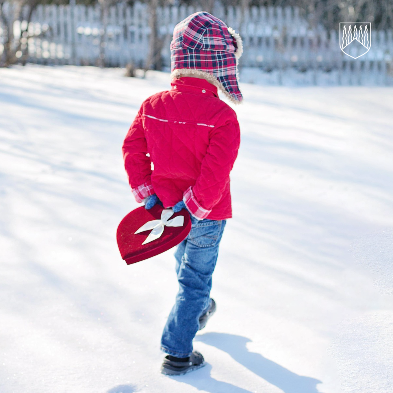 Lapsi punaisessa takissa kävelemässä hangella pidellen selän takanaan punaista sydämen muotoista rasiaa, jossa valkoinen rusetti.