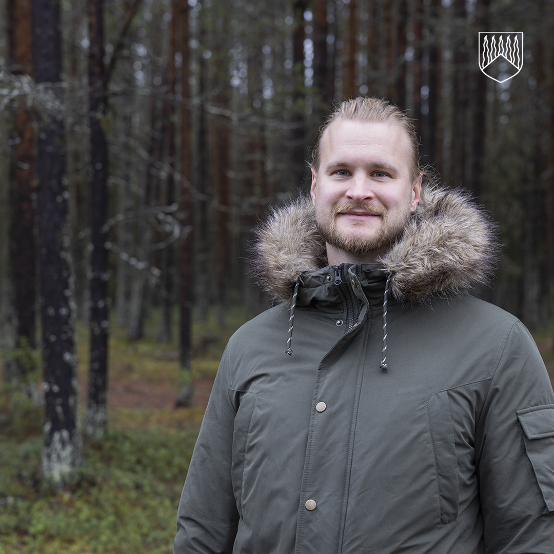 Henkilö seisoo metsässä, jossa taustalla näkyy tiheästi kasvavia, suoria mäntyjä ja sammalpeitteinen maaperä. Henkilöllä on yllään tummanvihreä talvitakki, jossa on suuri, karvareunuksinen huppu ja nappikiinnitys edessä. Takissa on myös vetoketju ja kiristysnyörit hupussa sekä tasku hihassa.