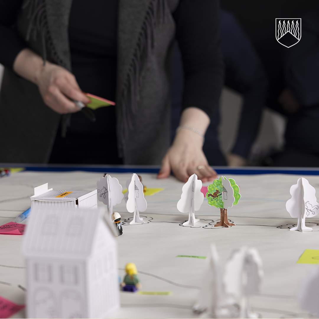 Henkilö työskentelee pöydän ääressä, jolla on paperille piirretty aluekartta ja sen päällä pieniä kartonkitaloja, paperisia puufiguureja ja värillisiä muistilappuja. Etualalla näkyy yksityiskohtaisia pienoismalleja rakennuksista ja puista.