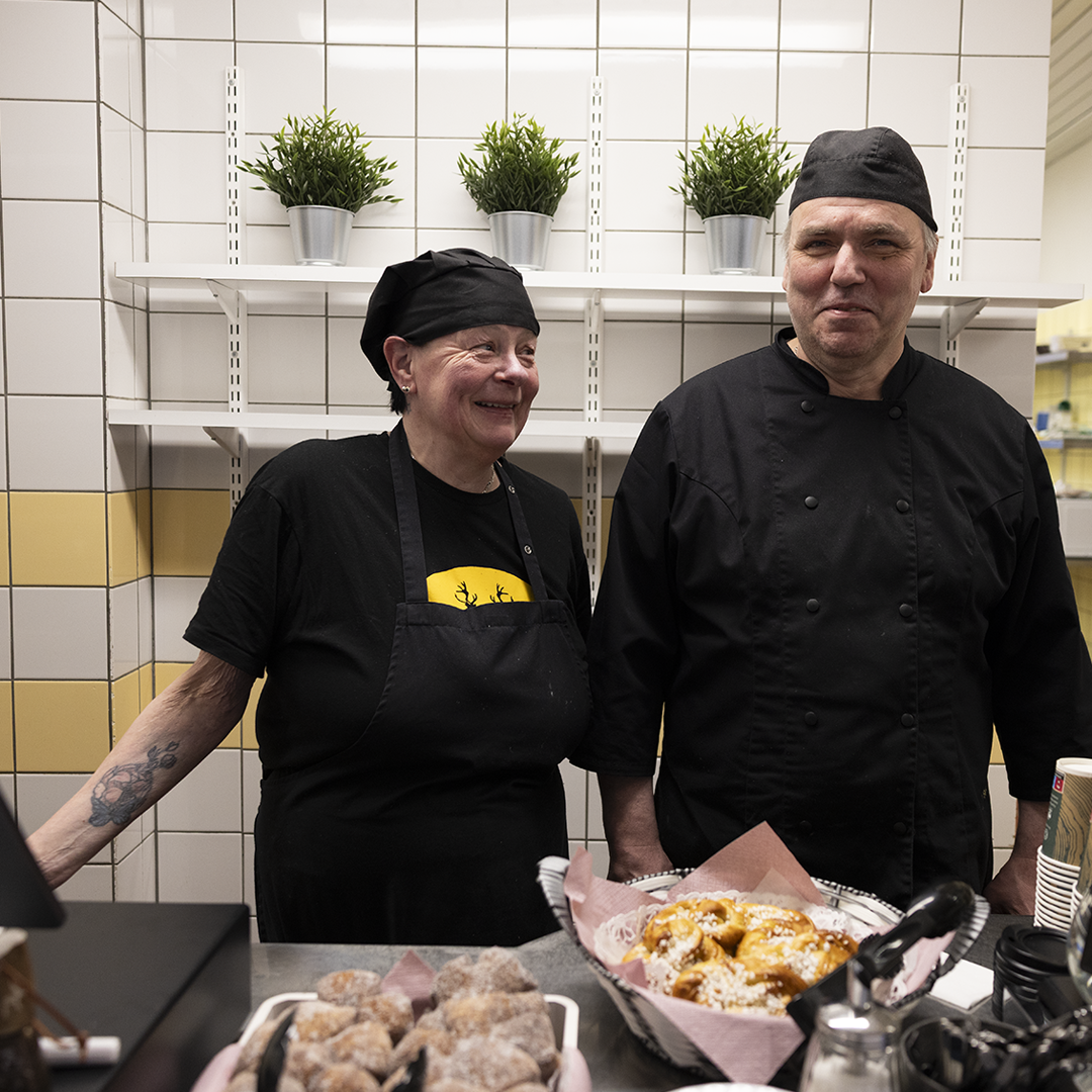 Kahvilan palvelutiski, jossa työntekijä annostelee ruokaa. Tiskillä on esillä leipiä, pullia, juomia ja muita tarjoiluja. Keskellä kuvaa näkyy ”Jaskan Kahvila” -logo.