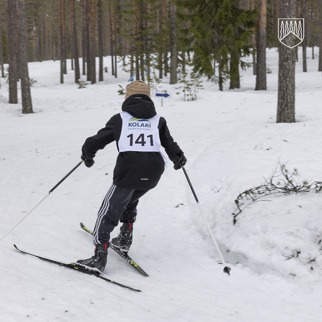 Hiihtäjä liikkuu latua pitkin lumisessa metsämaisemassa. Selässä on kilpailunumero 141 ja teksti ”Kolari”. Reitti kääntyy metsän keskellä, ja taustalla näkyy opaste nuolineen.
