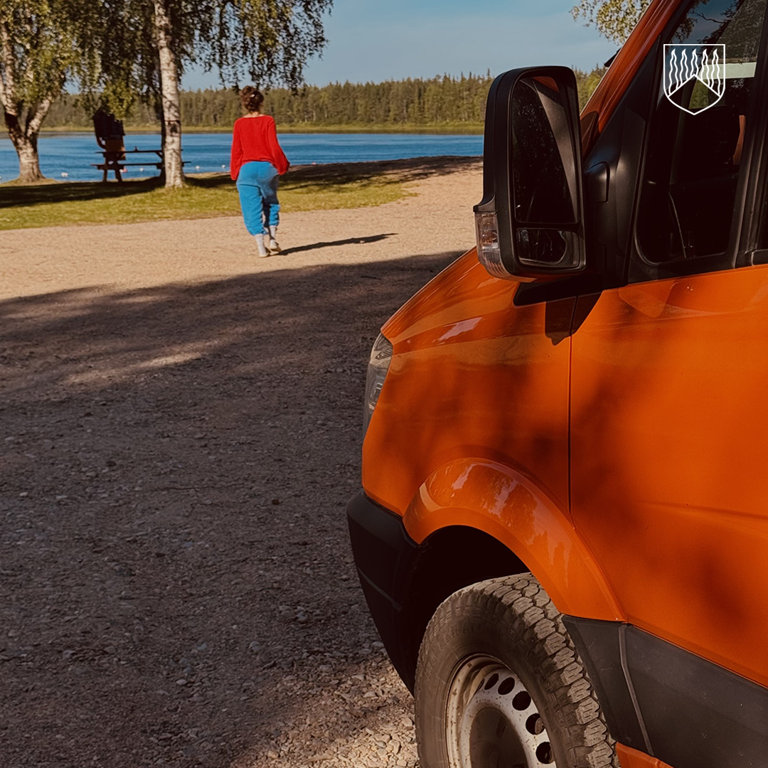 Oranssi matkailuauto on pysäköity sorapintaiselle matkaparkkialueelle järven rannalle. Taustalla henkilö kävelee kohti vesirajaa. Alueella kasvaa koivuja, rannassa näkyy penkki ja vastarannalla metsää aurinkoisessa syyssäässä.