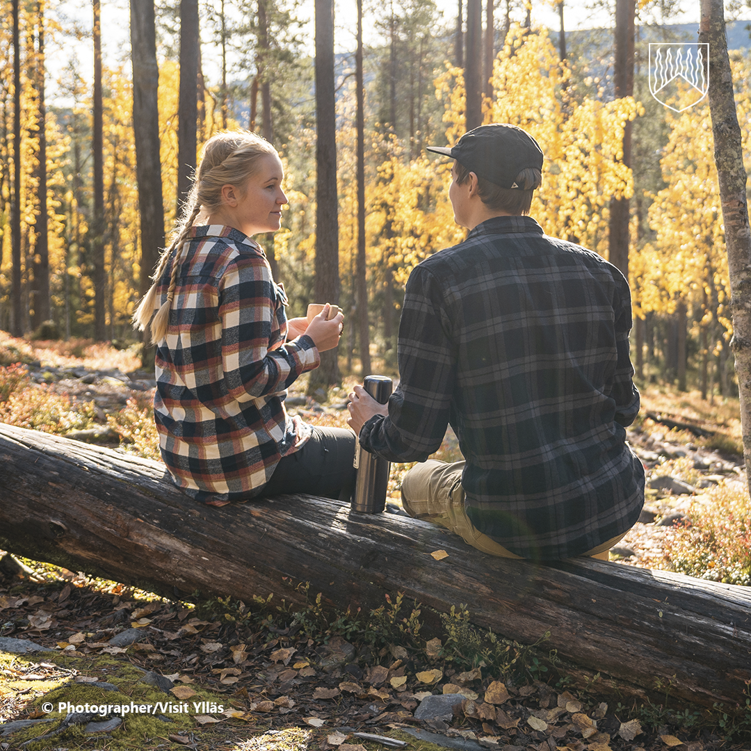 Kaksi retkeilijää istuu kaatuneen puun rungolla syksyisessä metsässä Ylläksen alueella. Taustalla näkyy keltaisena hehkuvaa ruskaa ja mäntymetsää. Toisella retkeilijällä on kädessään evästä ja toisella termospullo. Kuvan tunnelma korostaa luonnossa liikkumista, taukoilua ja virkistäytymistä reitistön varrella.