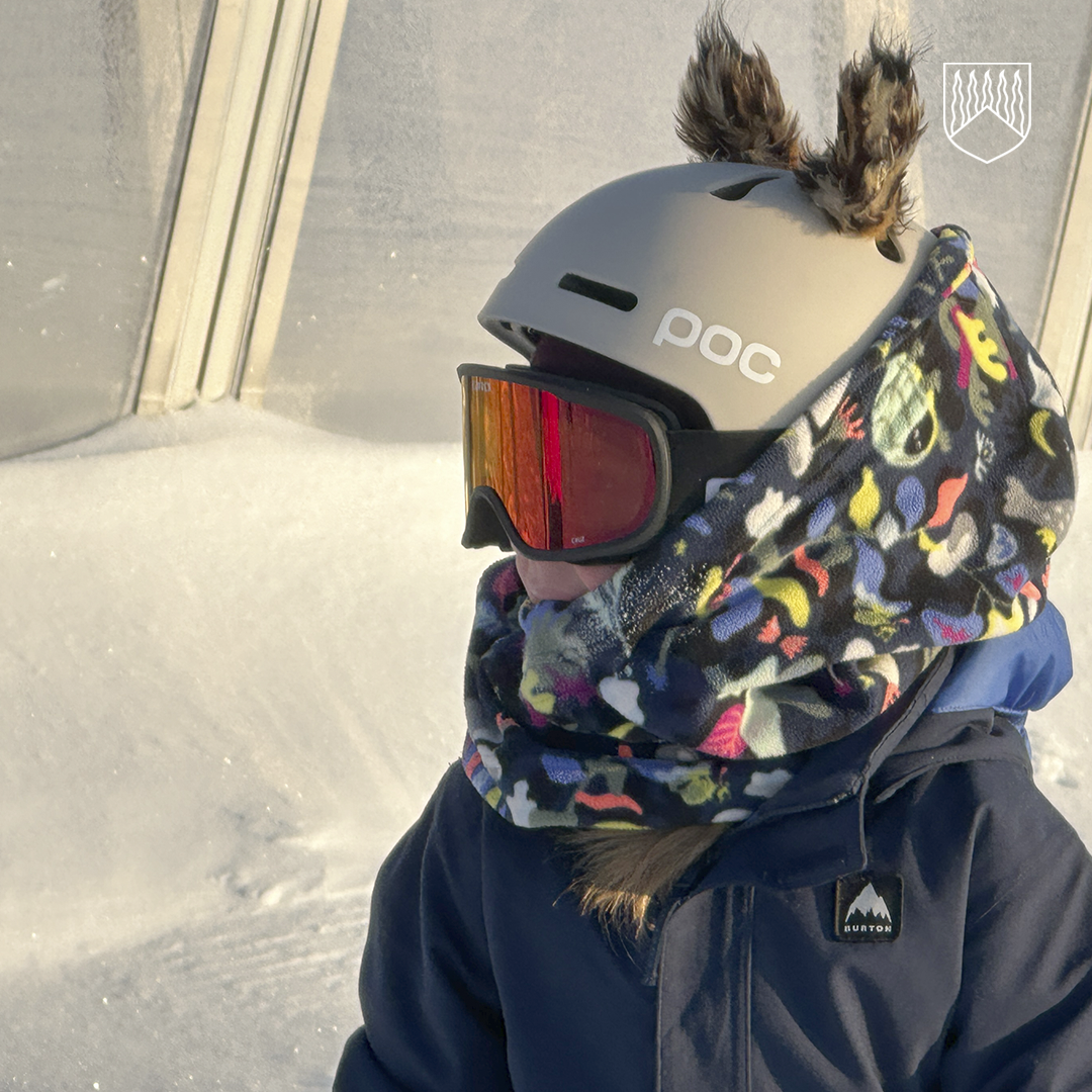 Lapsi seisoo talvisessa auringonpaisteessa laskettelukypärä ja punasävyiset laskettelulasit päässään. Kypärässä on pörröiset korvat, ja kaulaa suojaa värikuvioinen huivi. Taustalla näkyy luminen rinne ja vaalea rakenne.