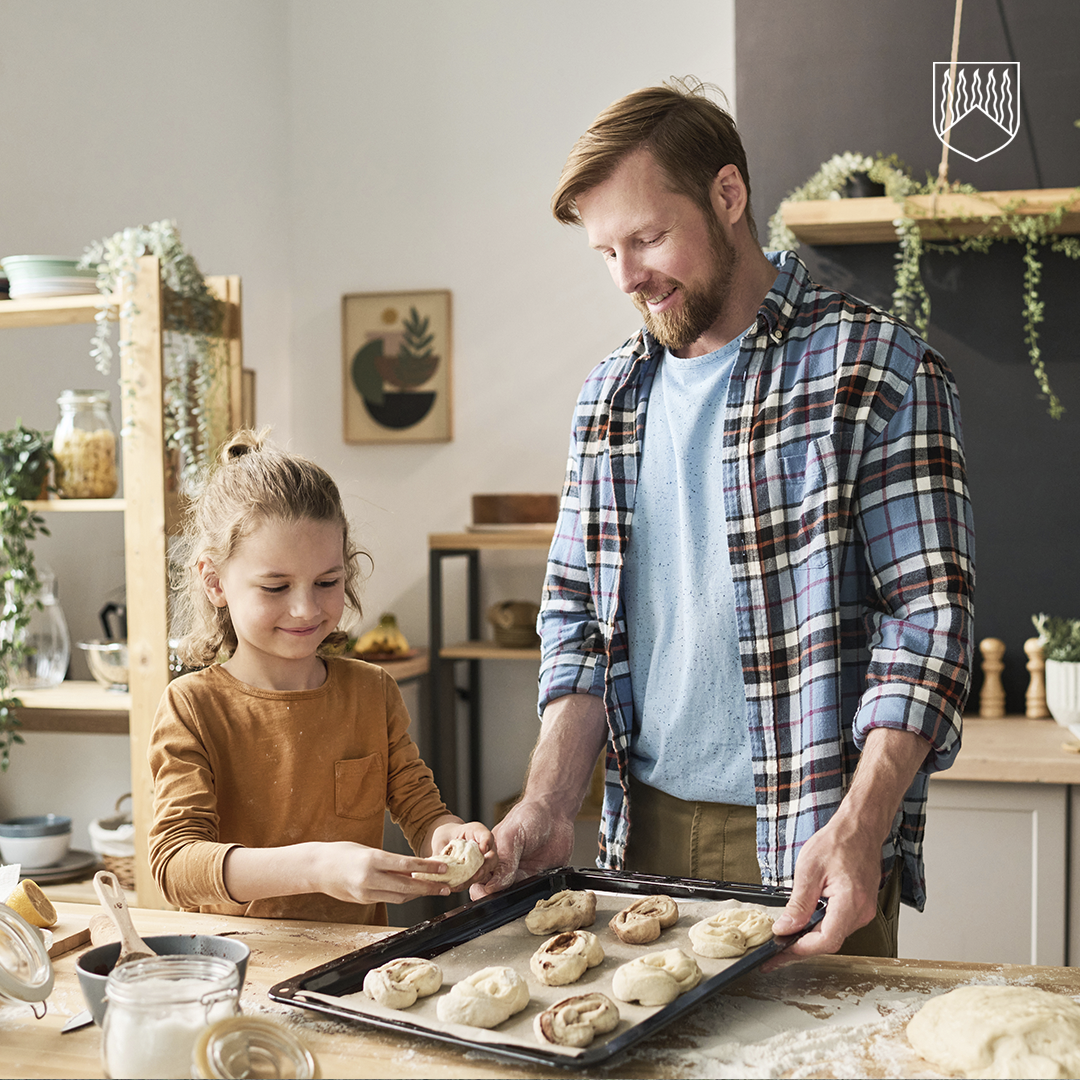 Lapsi ja aikuinen leipovat pullia yhdessä keittiössä. Uunipellillä on valmiiksi muotoiltuja pullia ja pöydällä leivontatarvikkeita.