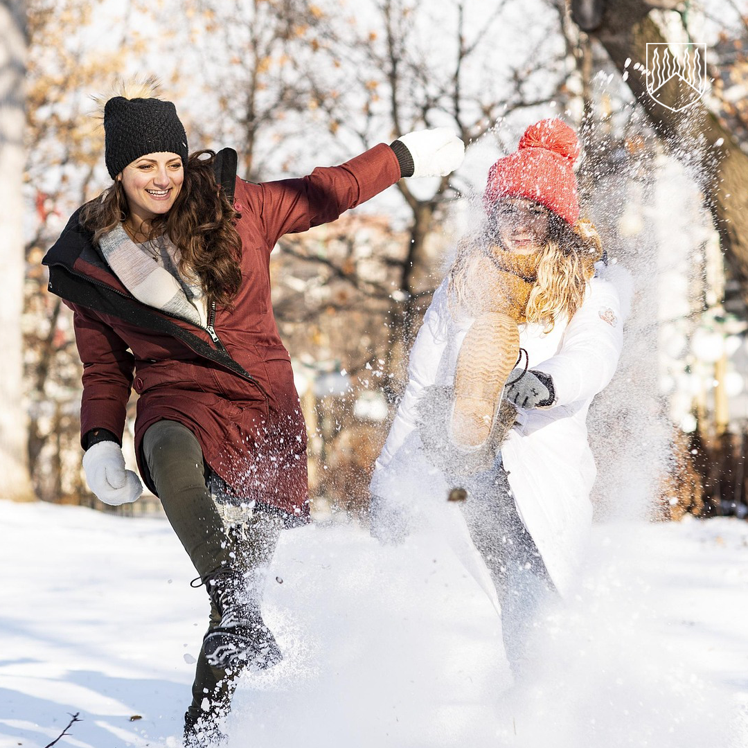 Kaksi henkilöä leikkii lumessa talvipäivänä potkien lunta ilmaan. Toinen on pukeutunut tummanpunaiseen talvitakkiin ja toinen valkoiseen takkiin sekä punaiseen pipoon. He näyttävät nauravan ja nauttivan talvisesta ulkoilusta puiden täyttämässä puistossa.
