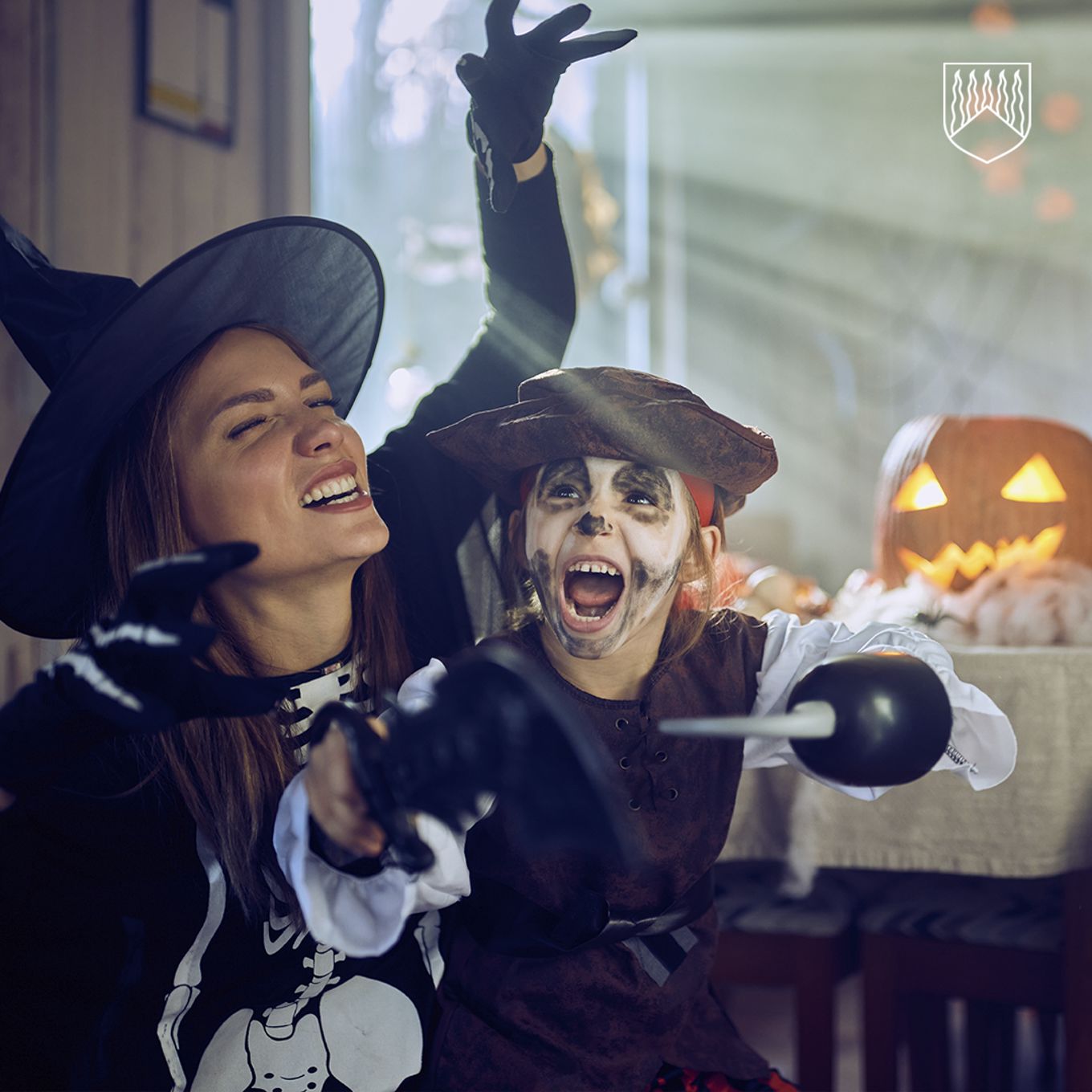 Kuvassa noitamaiseen halloween asuun pukeutunut naishenkilö sekä luuranko-merirosvoksi pukeutunut lapsi. Kuvan taustalla keittiön pöydälle perinteinen kurpitsa, jonka valaistu sisäpuolelta.