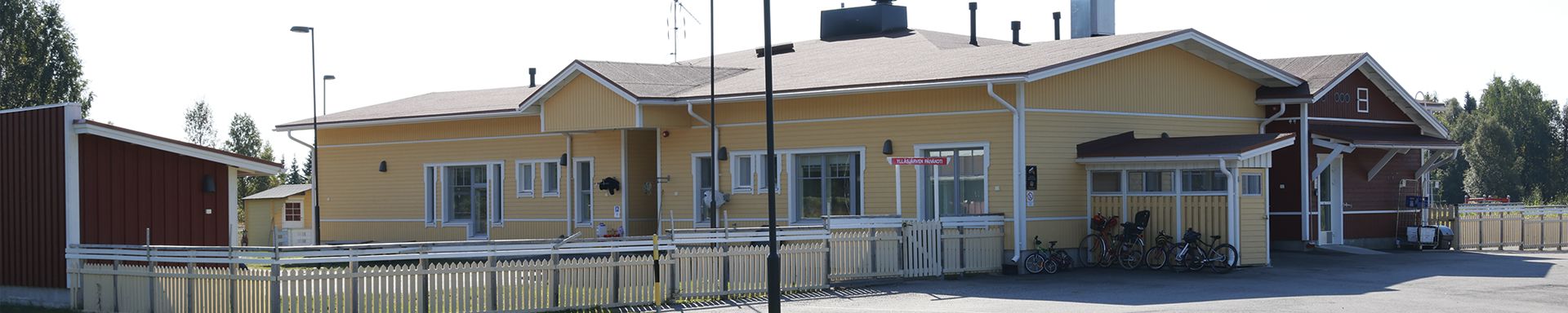 Yksikerroksinen keltainen rakennus, jossa on valkoiset reunukset ja useita ikkunoita sekä ovia. Oikealla puolella on pieni kuisti, ja oven vieressä punainen kyltti, jossa lukee 'Kunnantalo'. Rakennuksen edustalla on valkoinen aita ja useita polkupyöriä. Vasemmalla näkyy pienempi punainen rakennus ja taustalla puita.